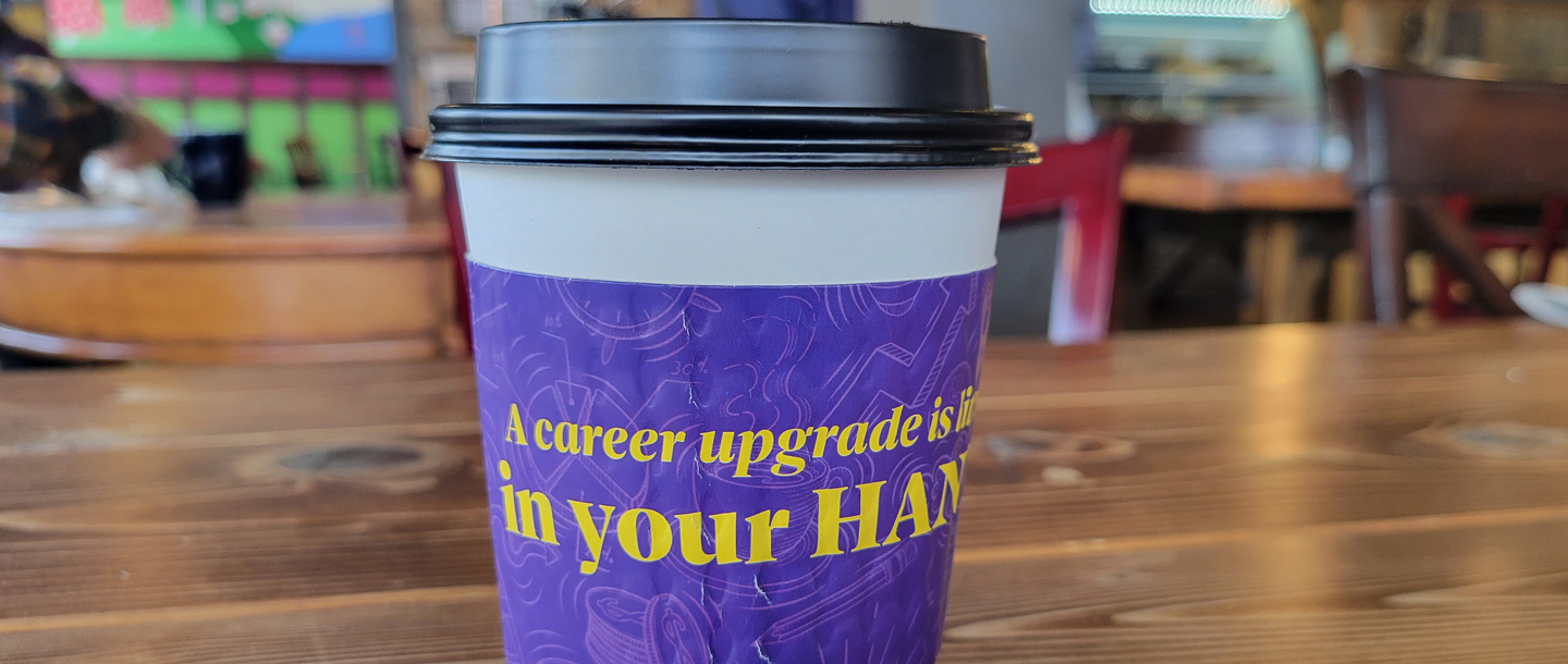 A coffee cup with lid and purple sleeve that has yellow text displayed on a table